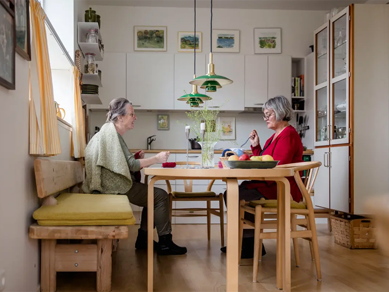 Two senior women eating at a wooden dining table with green pendant lights overhead