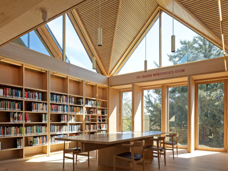 View of a second-storey reading room, showing a central table alongside a bookshelf and large window looking out over the trees.