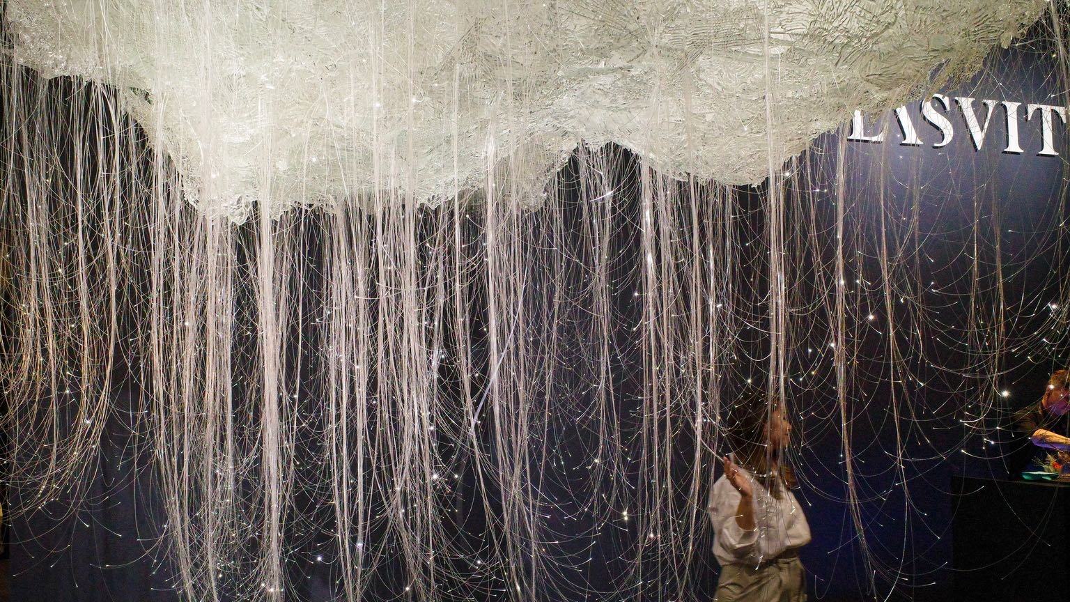 A person walks under a glass cloud with hanging light tubes at Lasvit's booth at Euroluce 2023.