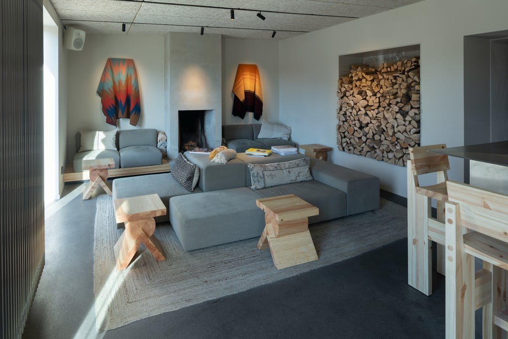 A series of low grey sofas and wood stools sit next to a wall niche lined with fire wood. A fireplace at the back of the room sits in between two sarapes hung on the wall.