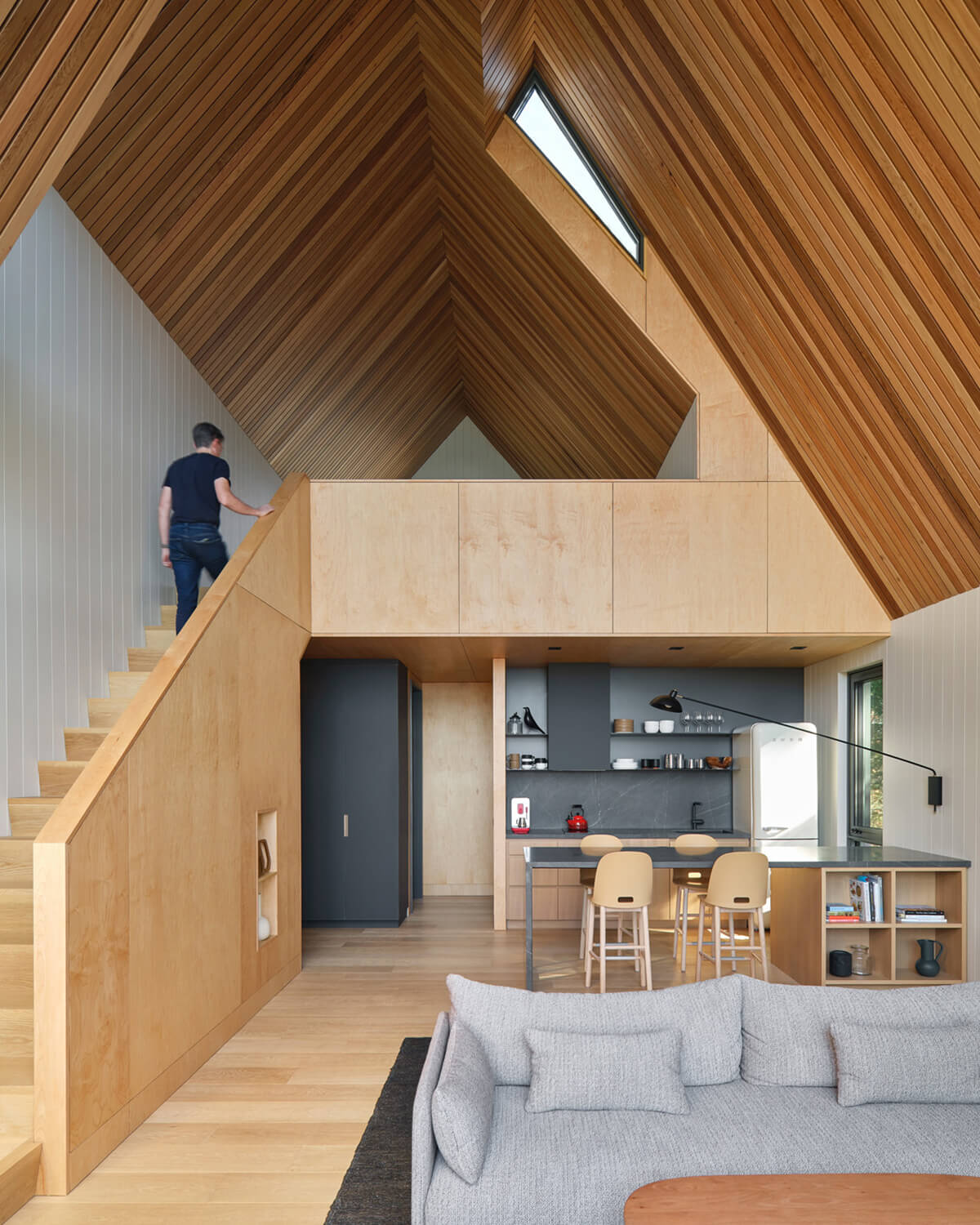 The western red cedar slat ceiling showcases the modern A-frame cabin’s angles. The compact kitchen with a backsplash and island in Greystone, a type of limestone, subtly contrasts the wood tones. The Alfi stools are from Emeco and the Waldorf sconce is by Lambert & Fils.