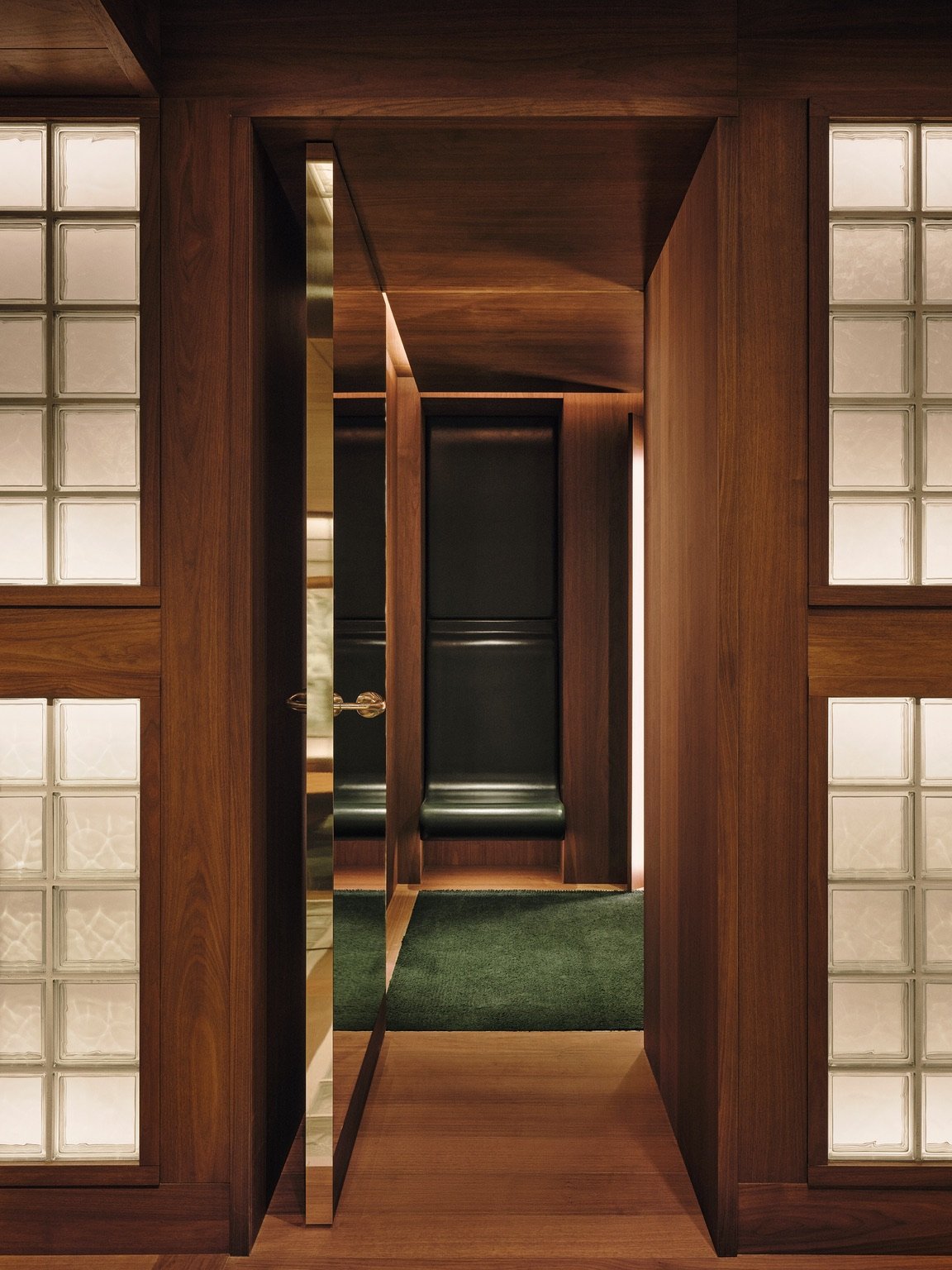 Glass block screens sit on either side of a doorway leading into a walnut-panelled room with a green rug.