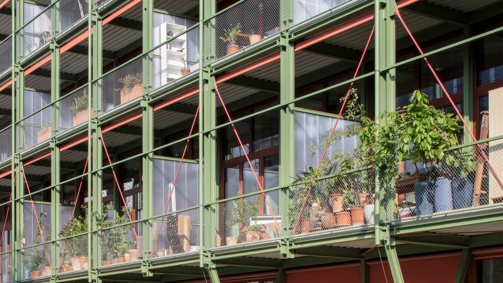 The southeast-facing façade of the new affordable apartment building in Basel, which was adapted from the Coop wine storage facility, expresses an eclectic character, with operable shades in terracotta and a light green metal frame system and railings.
