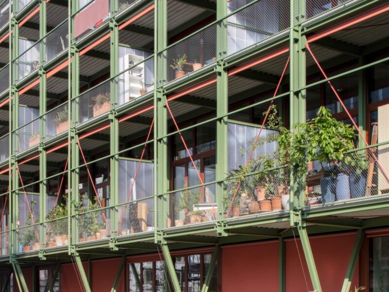 The southeast-facing façade of the new affordable apartment building in Basel, which was adapted from the Coop wine storage facility, expresses an eclectic character, with operable shades in terracotta and a light green metal frame system and railings.