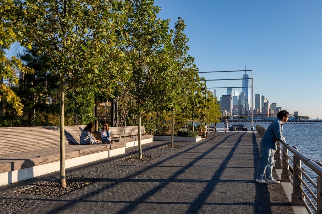 Two people sit on a wooden bench while a third leans on a railing looking out at the water at Gansevoort Peninsula Park in New York City.