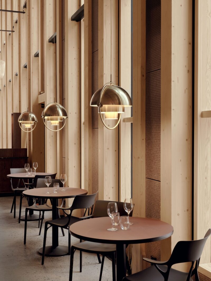 An interior view of Stockholm restaurant Rummel showing a row of black circular dining tables inside of tall, cathedral-like space lined in wood with hanging brass pendant lamps.