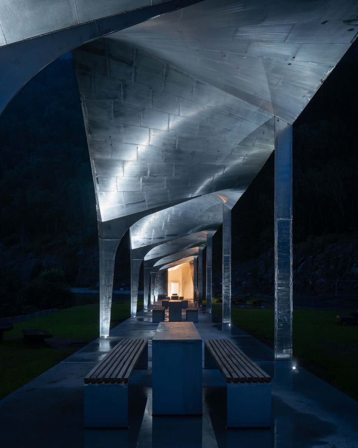 Looking towards the glowing washroom facility at the end of a roadside stop structure by Code Arkitektur that's illuminated at night. A picnic table sits in the foreground underneath the arched roof that shelters the walkway.