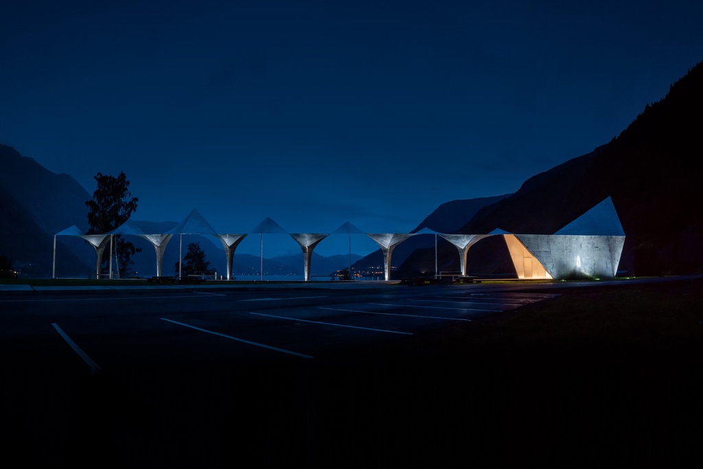 A night view of a roadside stop in Norway by Code Arkitektur. A rippling metal ceiling covers a narrow walkway that terminates in a glowing. metal washroom structure. The Hardangerfjord sits in the background.