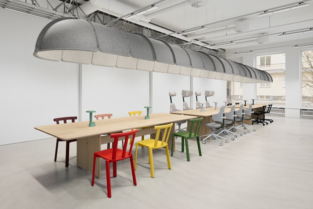 A row of new product launches — multicoloured dining and office chairs — sits at a long wooden table under a pill-shaped acoustic light in the office of Form Us With Love as part of an exhibition during Stockholm Design Week 2024.