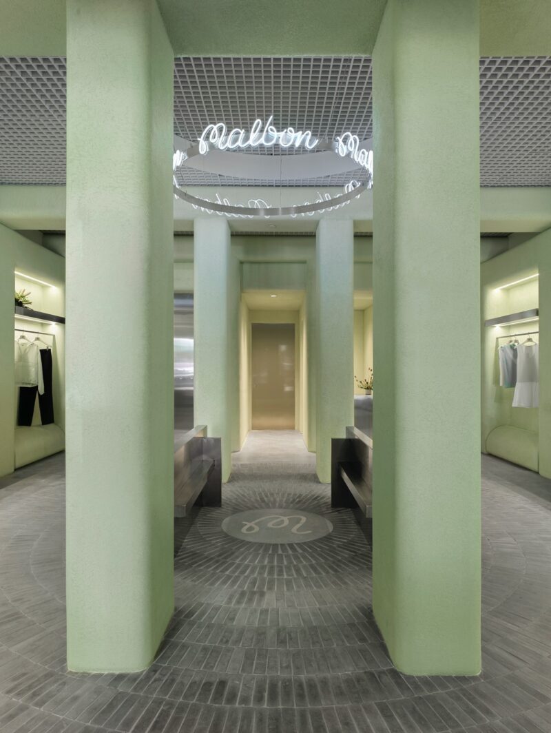 The centre of Malbon Golf Miami designed by 22RE, featuring four stuccoed pastel green columns. An aluminum ring hangs in the middle with the store's name written out in cursive neon.