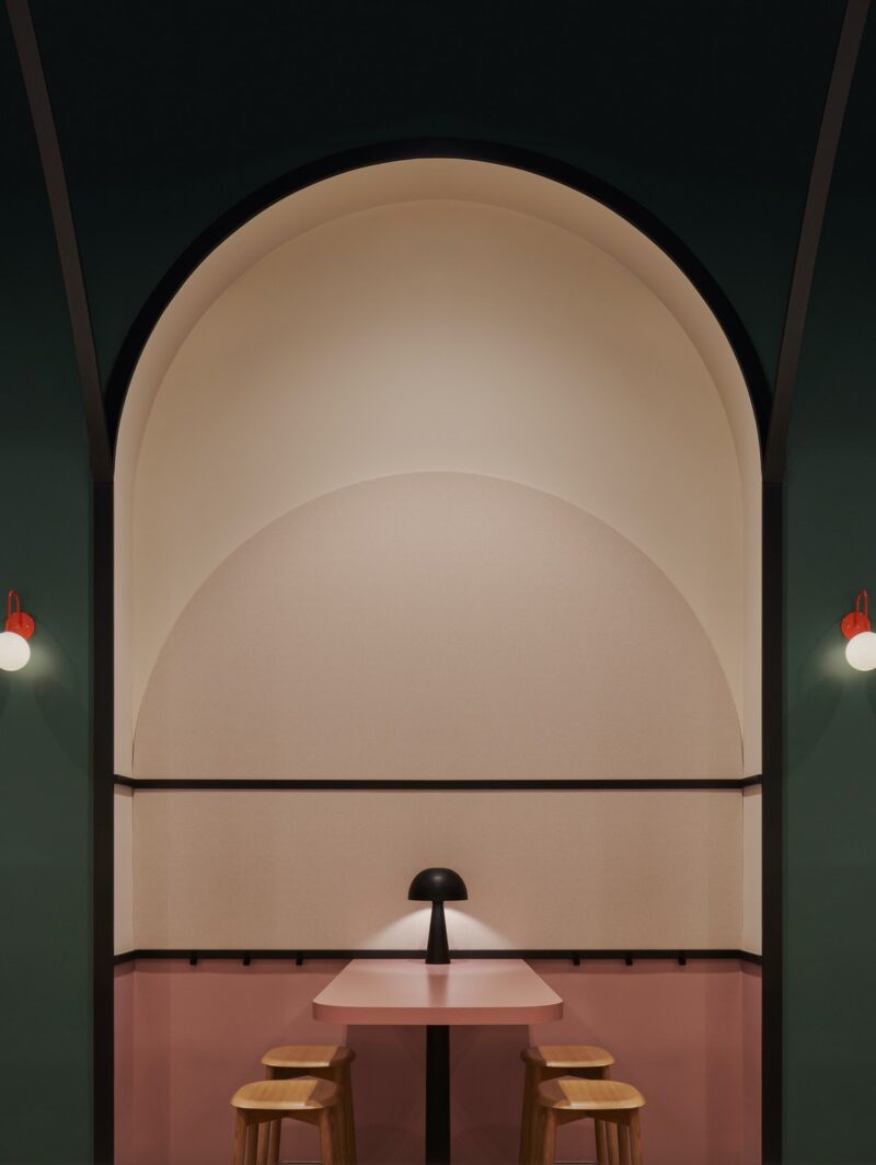 A curved wall niche at Talea Bryant Park, framed in green and featuring a soft mauve partway up and a cream paint for the top half of the arch. Four wooden stools sit around a pink bar table.