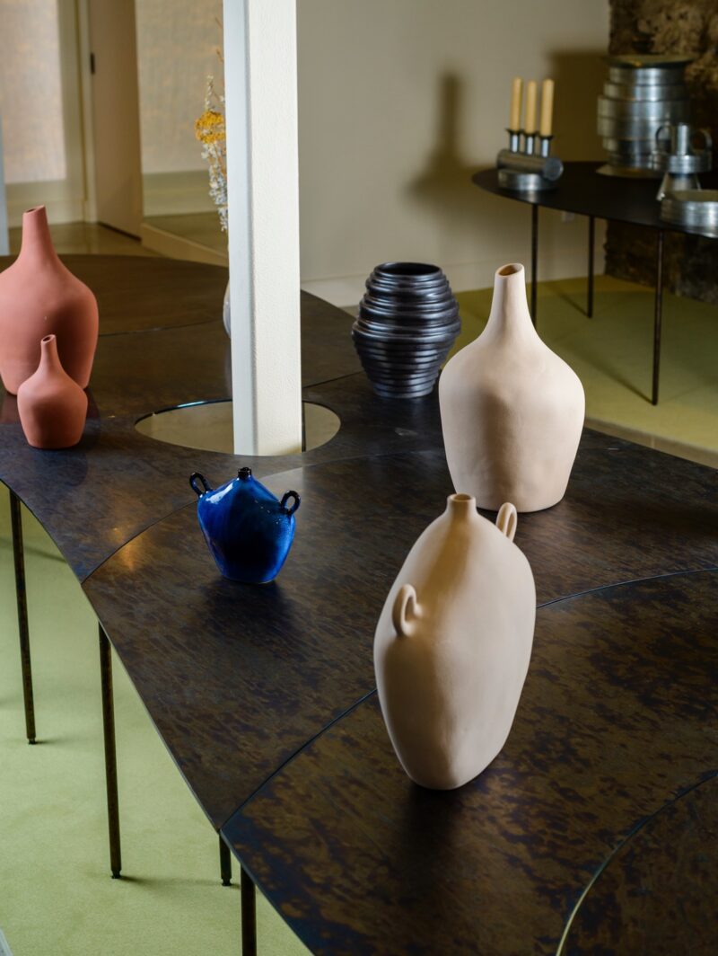 A selection of sculptural vases sit on a rounded black steel table in front of a natural stone wall at the Montreal store 207 Ouest. A circular cutout in the middle of the table allows a column to pass through.