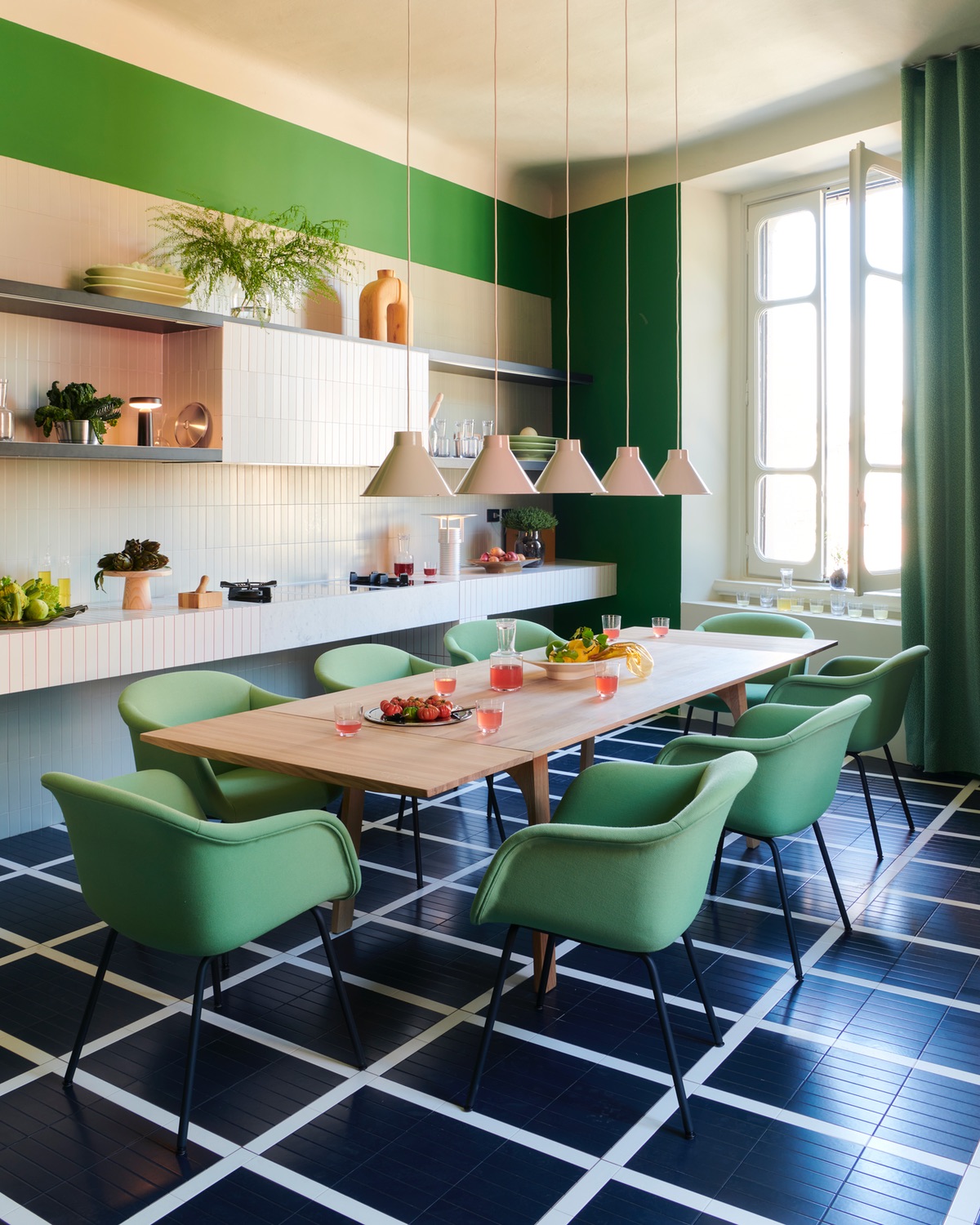 A wooden dining table surrounded by green chairs with beige pendants hanging above. A tiled white kitchen is to the left and the floor is covered in large black tiles with thin white outlines. Part of a Milan design week 2024 review that speaks to the value of quiet domestic spaces.
