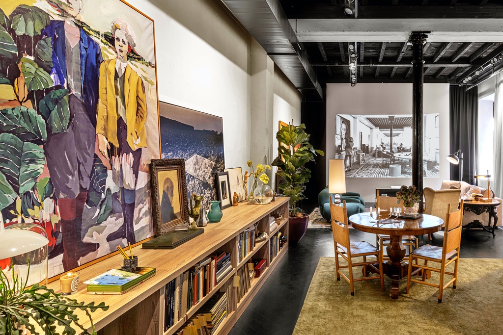 A wooden bookcase runs along the wall of a living room setup, with a large painted canvas resting on top showing a man in a blue suit and a woman in a yellow jacket. A round wooden dining table surrounded by chairs sits to the side, while various antique trinkets are scattered throughout. Part of a Milan design week 2024 review that speaks to the value of quiet domestic spaces.