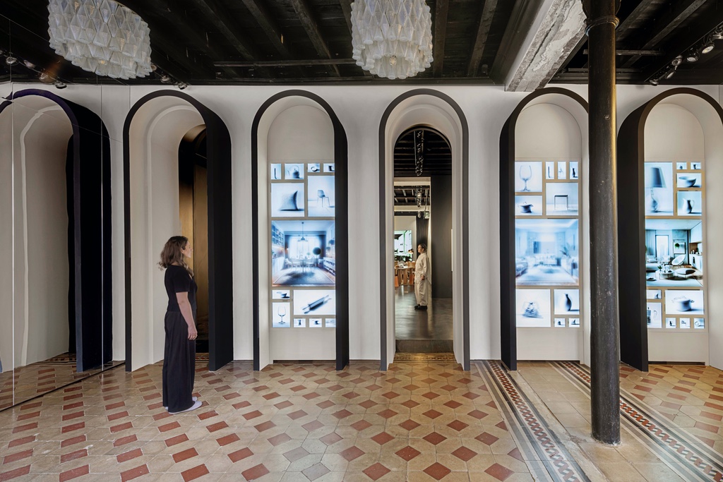 A woman stands in front of a row of black archways. Two of them feature several rows of screens, while one acts as an entrance into another room. The floor features a diamond terrazzo pattern.