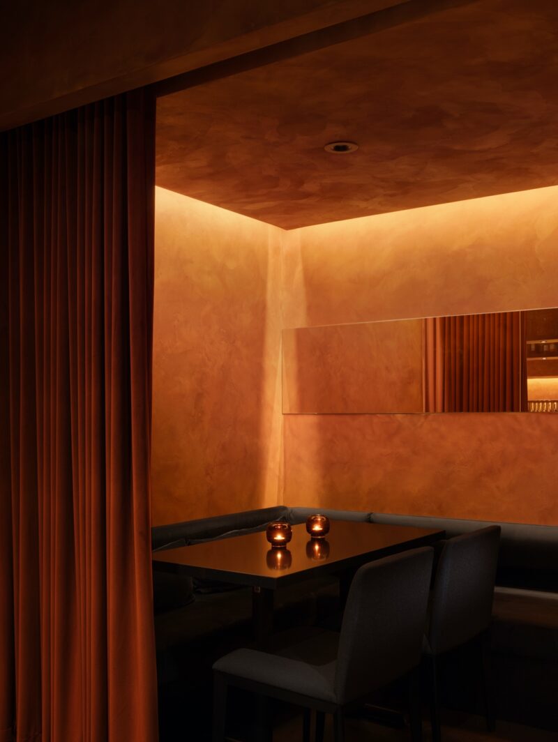 Ama restaurant by Vancouver design studio &Daughters is modeled after Blade Runner 2049. A black table sits in a dining nook closed off with orange velvet curtains. Behind it are a golden mirror and venetian plaster walls soaked in a golden glow.