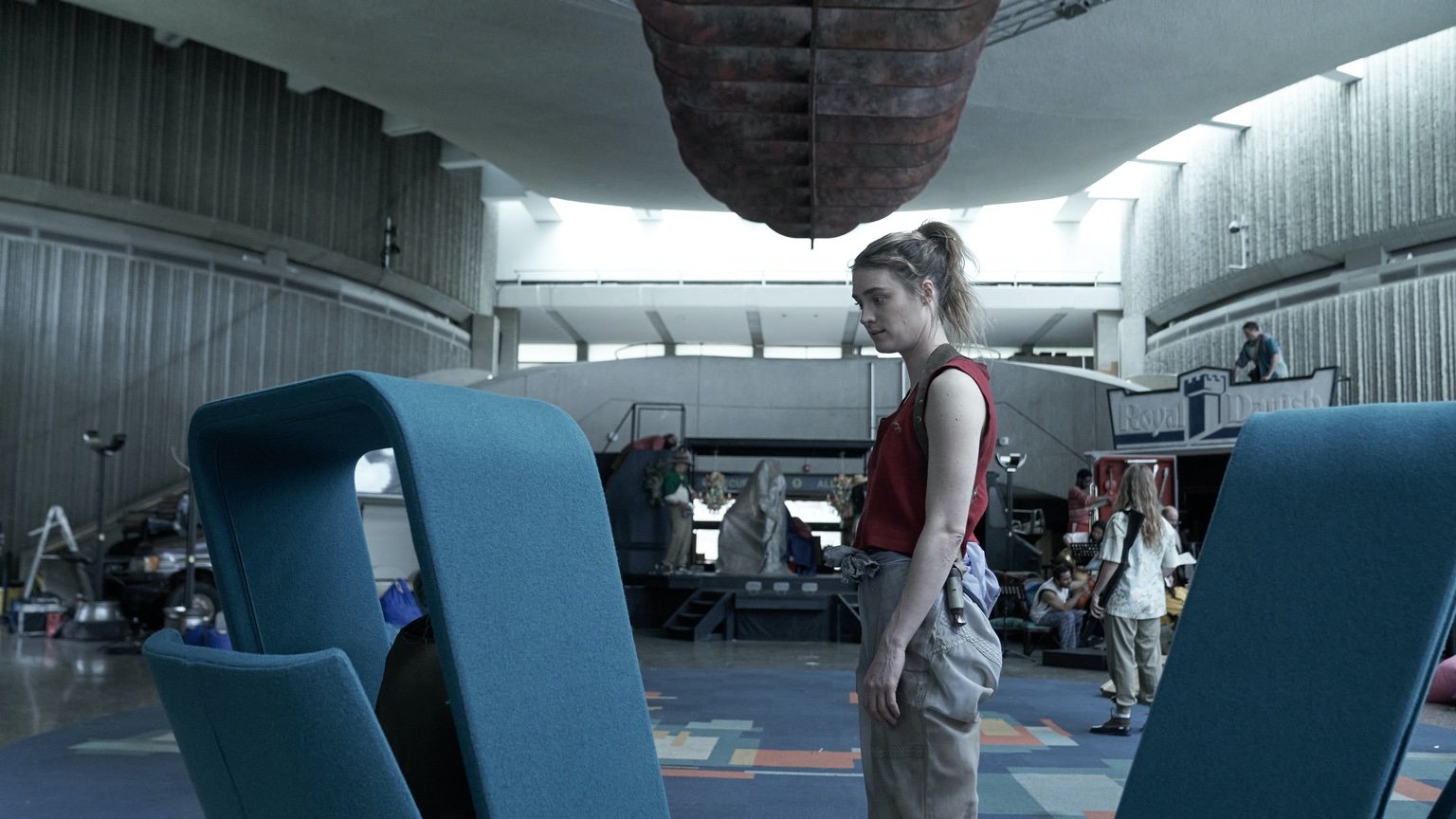 The actor Mackenzie Davis, a blonde woman with her hair in a pony tail, stands wearing a red tank top in between two modern blue chairs inside Severn City Airport (filmed in the brutalist Ontario Science Centre) in a still from the television show Station Eleven.