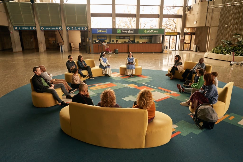 A group of people sit on yellow sofas gathered around a blue rug with a geometric pattern of the Great Lakes inside Severn City Airport (played by the brutalist Ontario Science Centre) in a still from the television show Station Eleven.