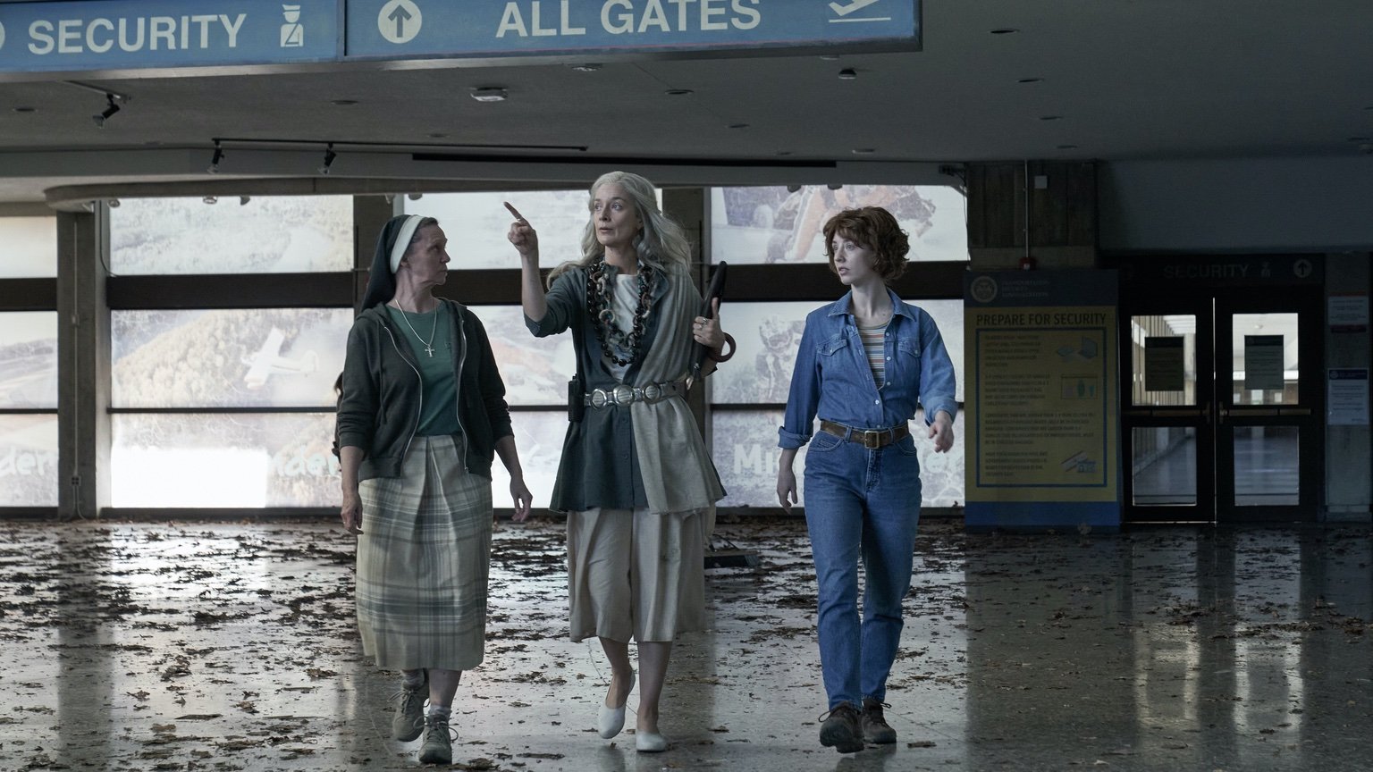 A woman with white hair walks through a worn-down version of Severn City Airport (played by the Ontario Science Centre) with a nun on the left side of her and a woman with curly hair in a blue jumpsuit on the right. Signs above say "Security" and "All Gates."
