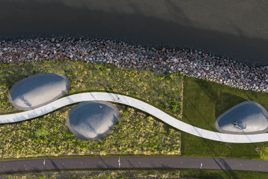 An aerial view of a snaking path with large pebble sculptures on either side. Grass separates the path from the St. Lawrence River.