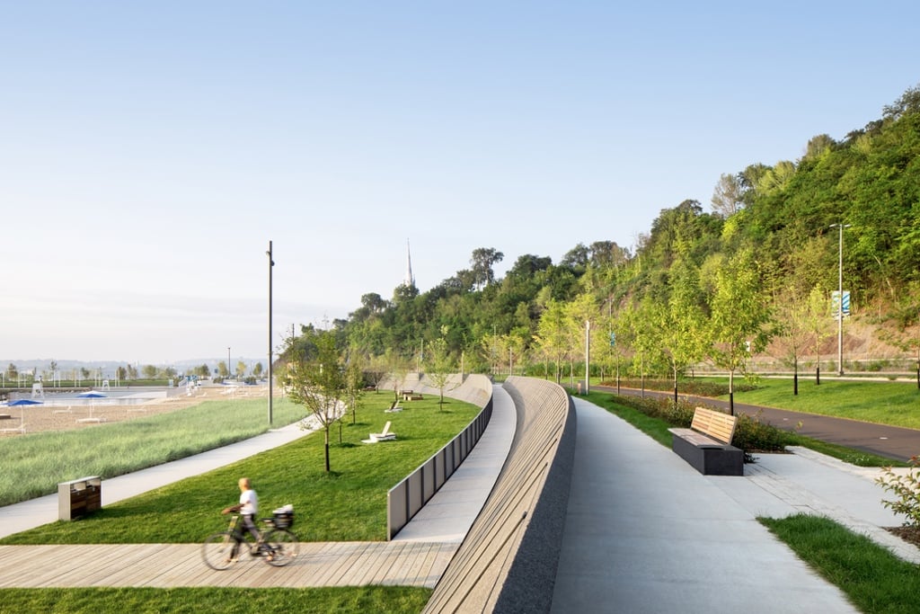 A biker moves along a wooden path perpendicular to a series of paved paths with a mountain range in the background.