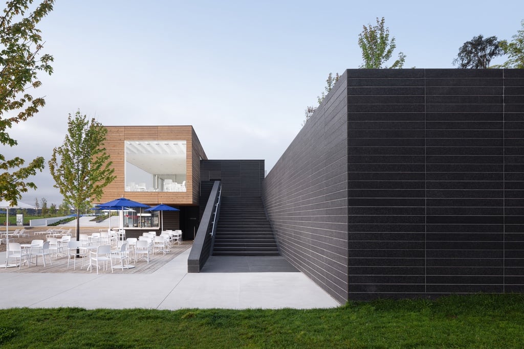 The pavilion next to the beach at Promenade Samuel-de Champlain features a staircase leading up to a cantilevered wood box with a big square window. Patio chairs sit underneath below blue umbrellas. To the right is a big brick wall.