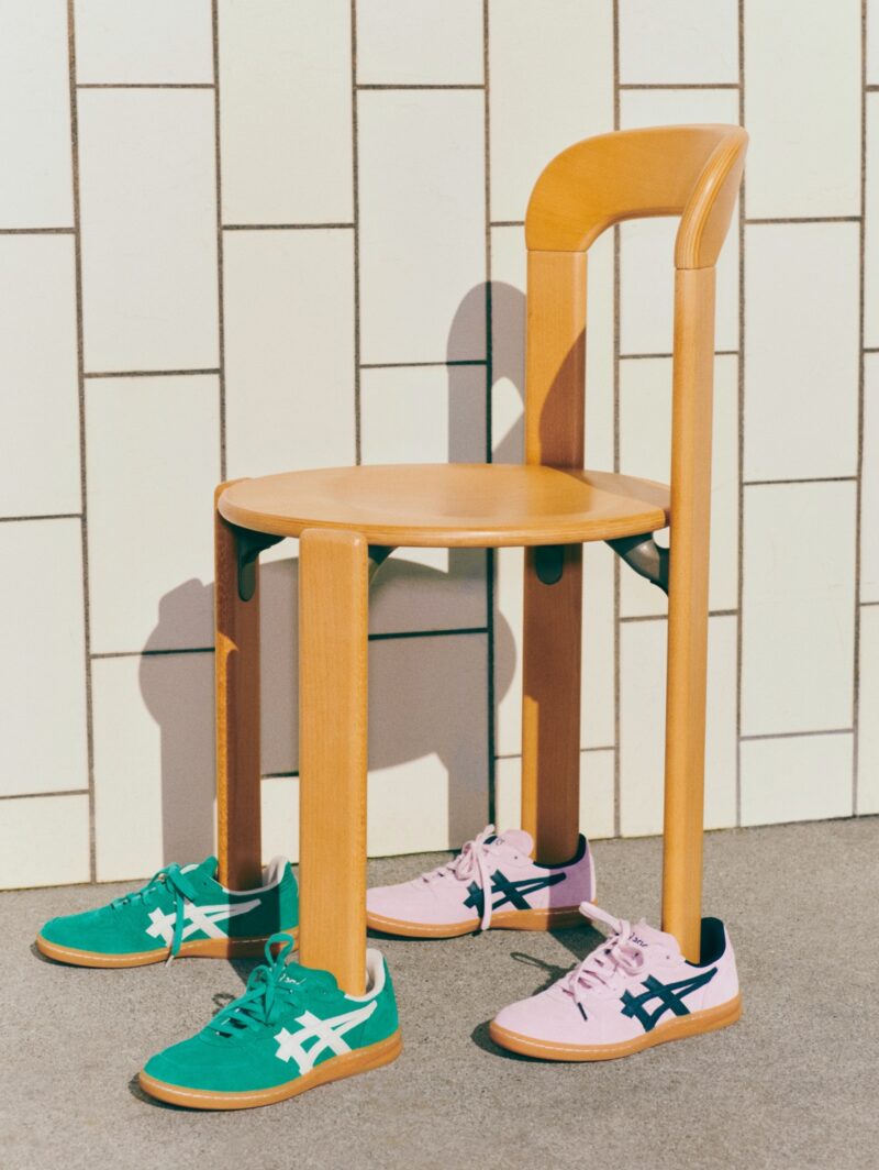 Two pairs of Asics sneakers (one green with white stripes, one pink with dark blue stripes) made in collaboration with design brand Hay, with the legs of a wooden Hay chair resting inside of them.