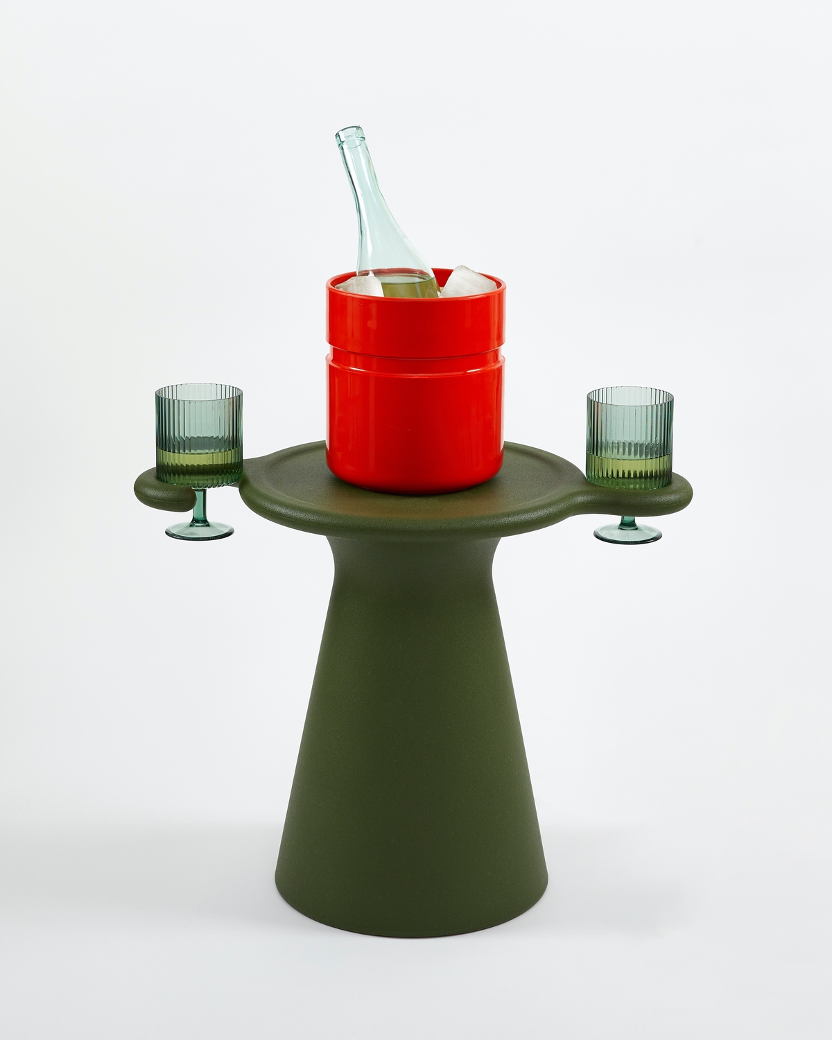 A green side table with a red ice bucket on top and two glasses resting inside the curled hooks on either side.