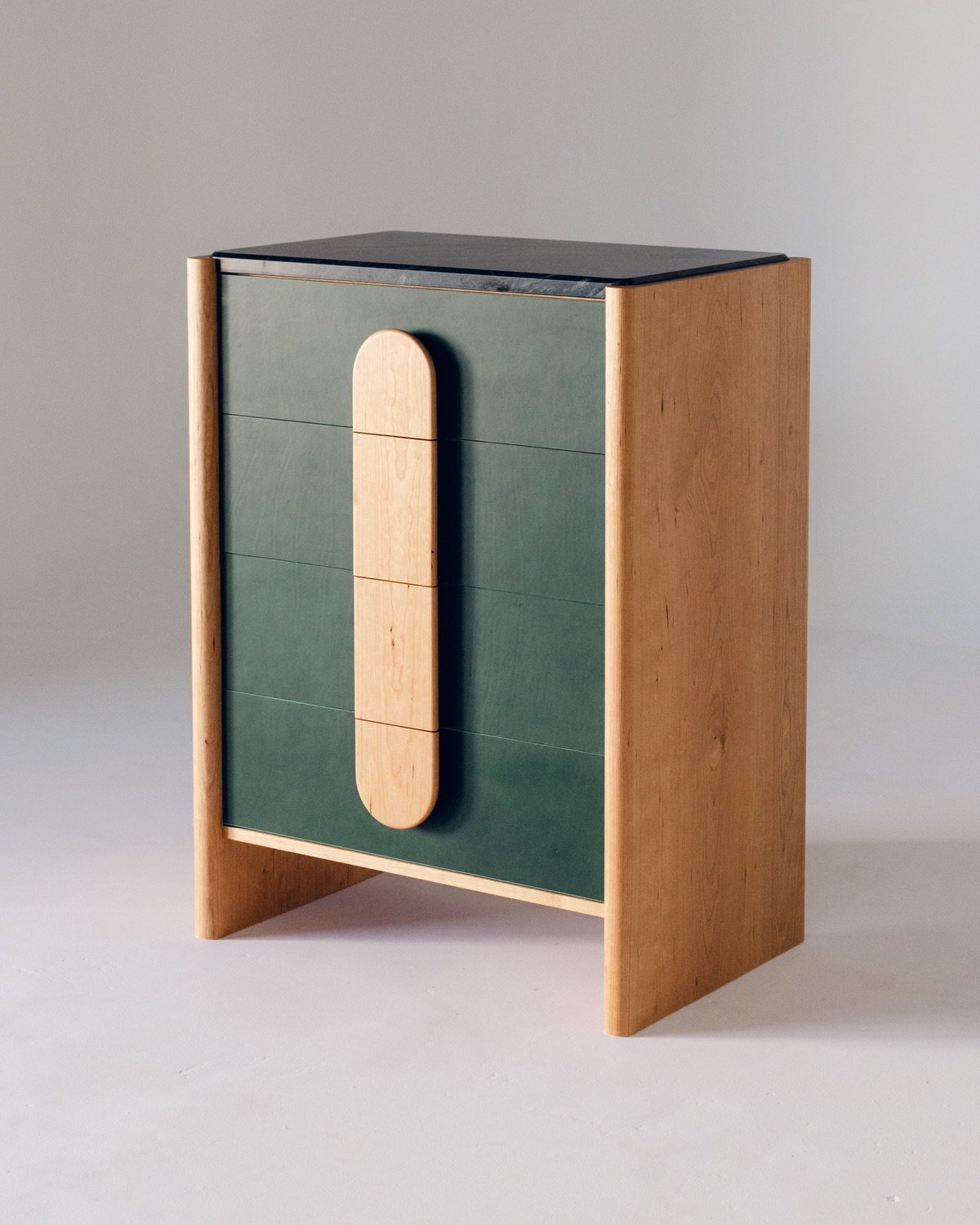 A chest of drawers with a green front, black marble top and wooden sides. The central handles for each shelf line up to form a pill shape.