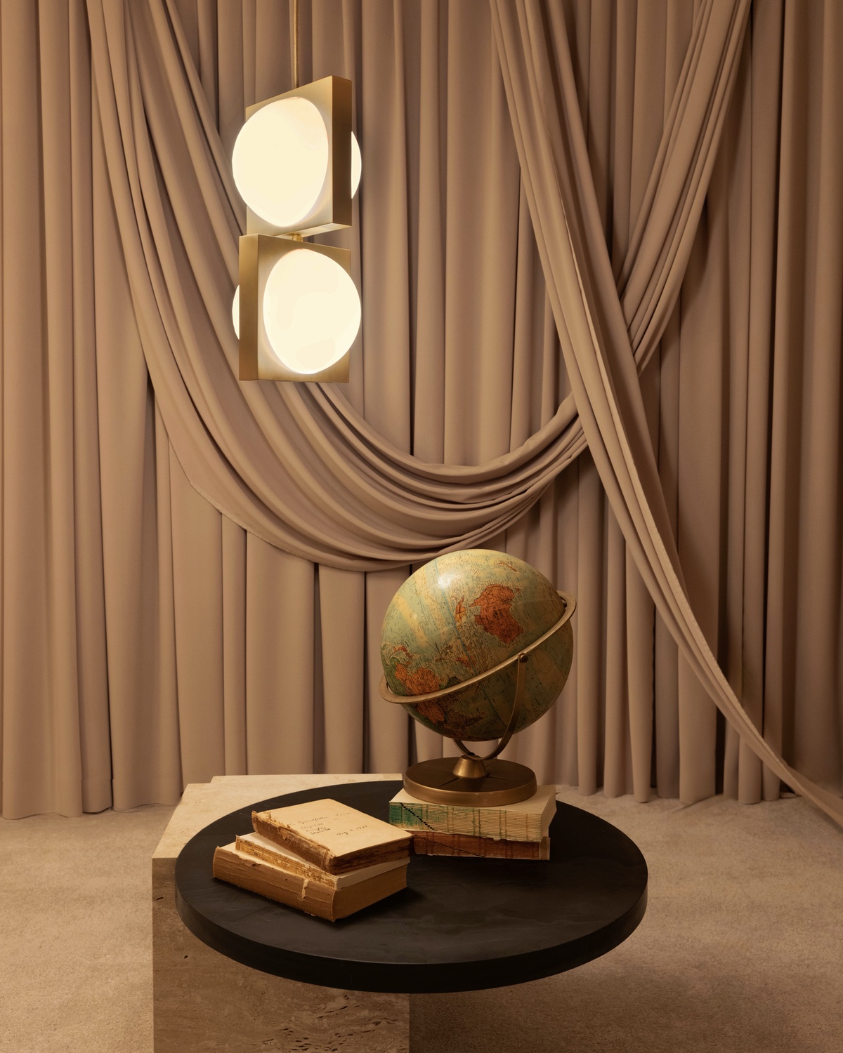 A Solstice pendantfrom the Alchemist collection by Lee Broom with glowing orbs inside of square brass frames. It hangs above a globe sitting on a table with a stack of books in front of a backdrop of a pink curtain.