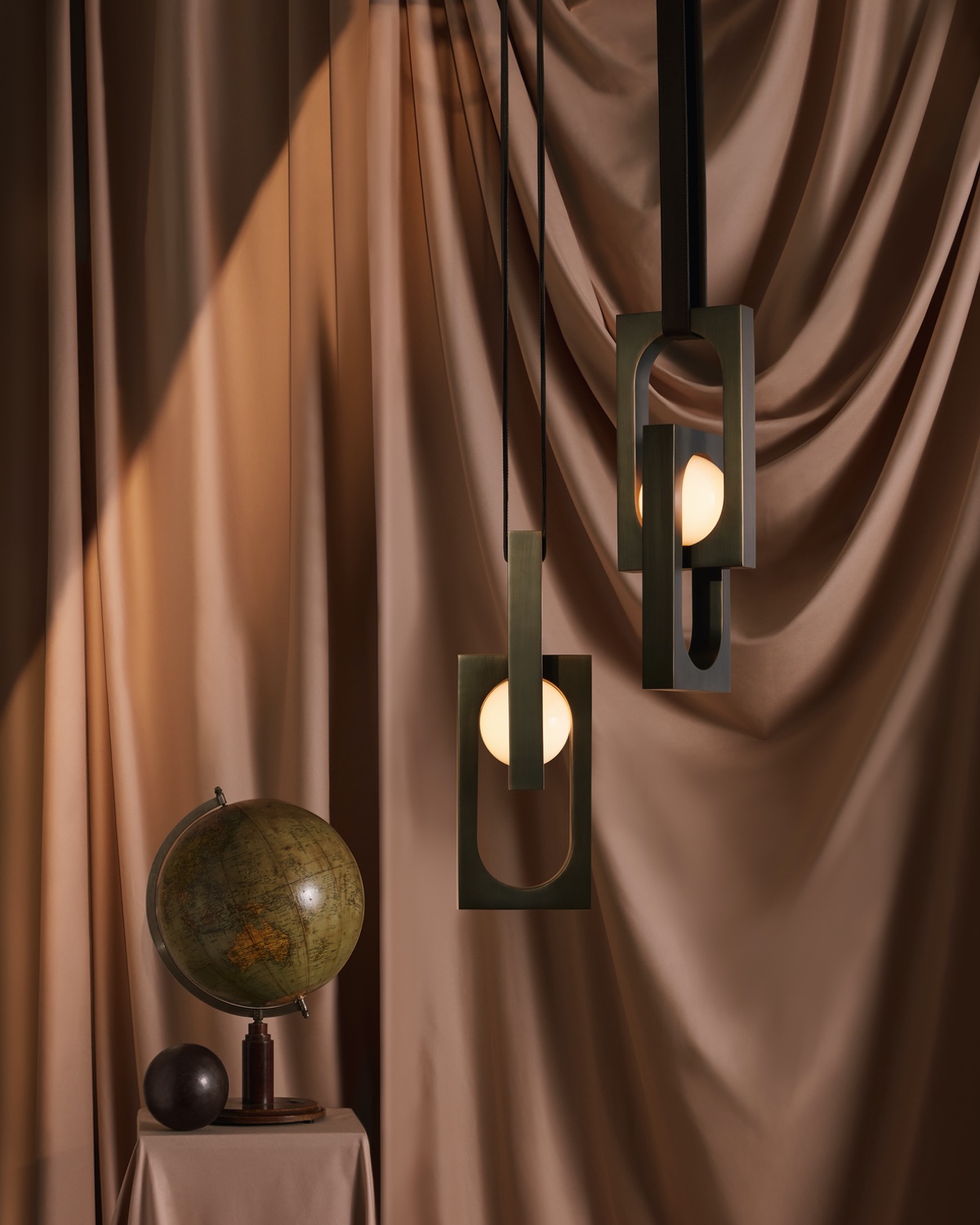 Two Mythos pendants, each with a glowing orb suspended inside of a rectangular brass frame that hangs from a leather strap.