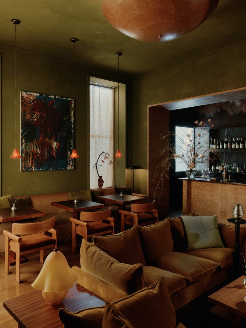 The bar room at Quarters in New York featuring a velvet sofa and three cocktail tables with wooden tables and glowing orange glass pendants hanging above. The room is painted moss green, and to the right is a burled wood bar table.