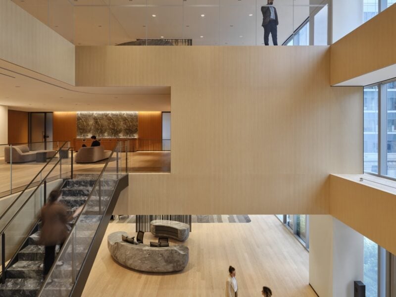 A view of an open, three-storey space in the Scotiabank North office designed by KPMB Architects in Toronto with light wood flooring and a marble staircase connecting the first two floors. A man stands in a glass window looking into the atrium from the third floor.