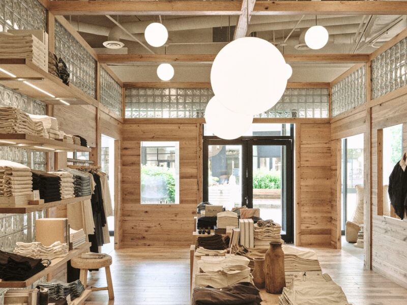 An image of a Buck Mason store design in Oak Brook, Illinois by Norman Kelley, featuring an oakwood shell structure built inside of a storefront. The top of the wooden structure is lined in glass brick and orb-style lights hang above tables of clothing in khaki and grey tones.