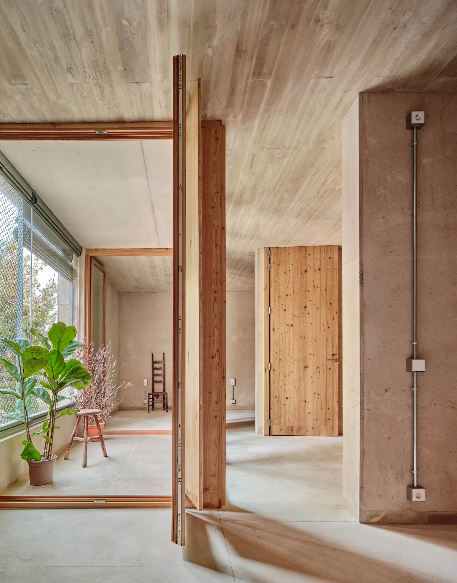 At the rear of a residential unit at Son Servera, the winter garden faces a capacious window with roll-up blinds. This indoor–outdoor space funnels passive energy into the home. Raw concrete and warm wood are the main interior materials.
