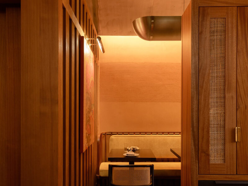 A long brass bowl-shaped pendant hangs above upholstered banquette seating in a gold-toned wooden space at Sway Aspen, a Thai restaurant designed by Michael Hsu Architecture Office.