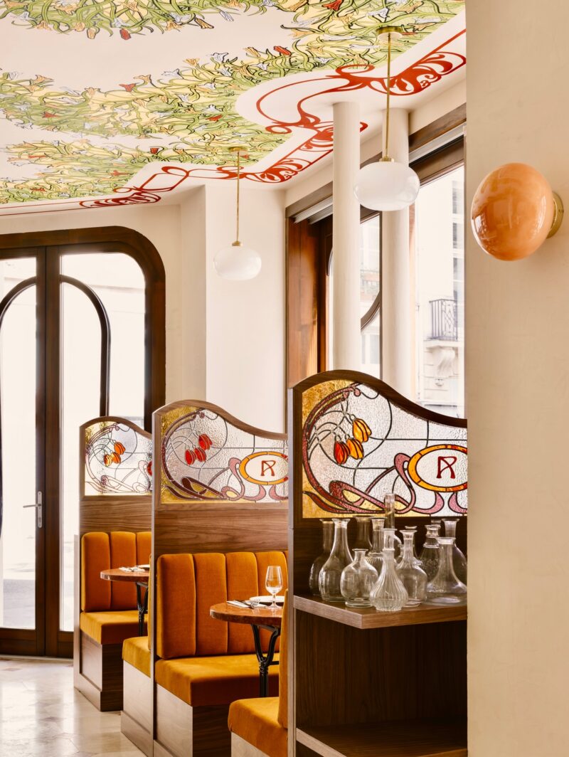 The booths at Adela pasta restaurant in Paris designed by Jessica Mille Architecte with stained glass dividers in between booths with orange velvet upholstery. The ceiling is painted with an elaborate floral mural.