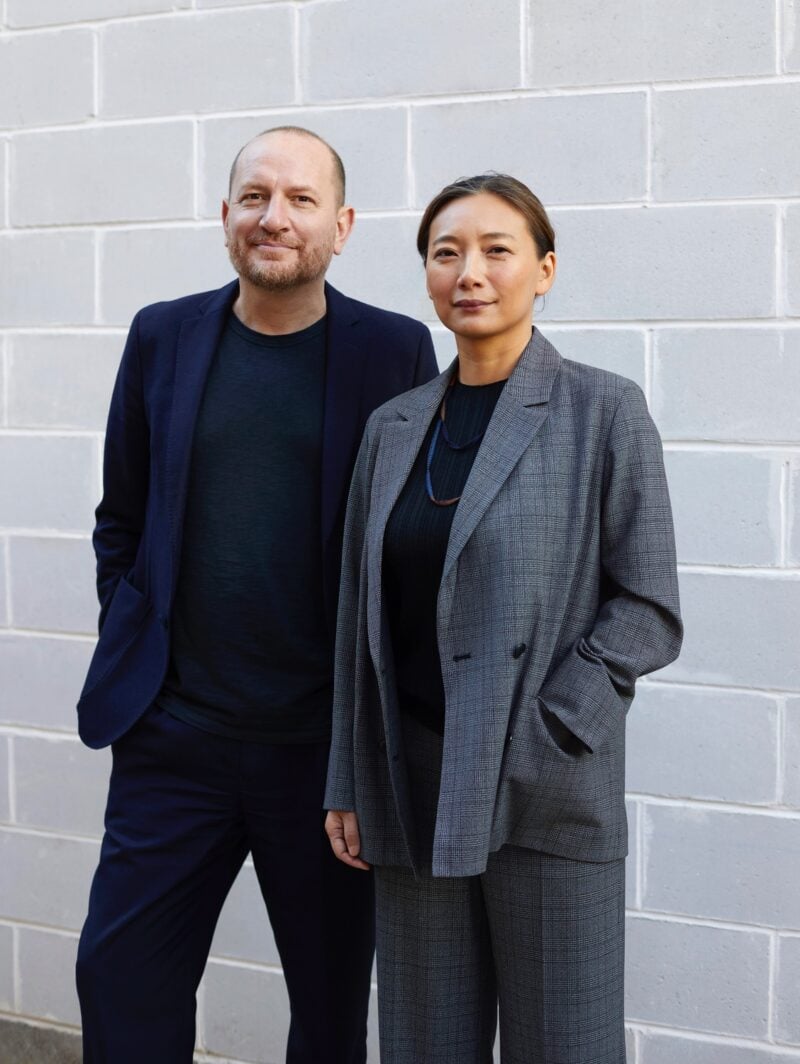 A portrait of the two principals that lead Brooklyn architecture studio SO – IL: Florian Idenburg (left, in a black suit) and Jing Liu (right, in a grey suit). Both stand in front of a grey brick wall.