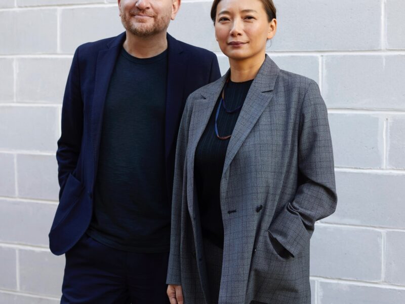 A portrait of the two principals that lead Brooklyn architecture studio SO – IL: Florian Idenburg (left, in a black suit) and Jing Liu (right, in a grey suit). Both stand in front of a grey brick wall.