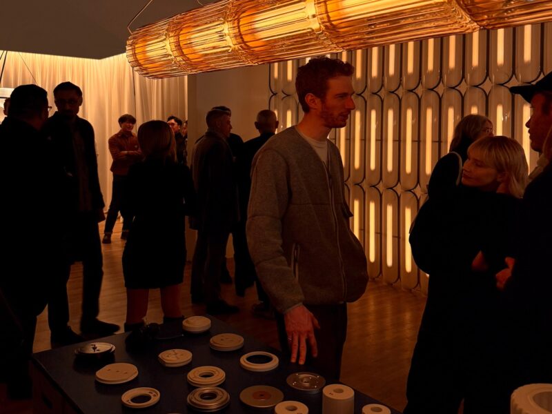 A group of people standing in a dark room illuminated by a horizontal tubular pendant shaped like a Grecian column.