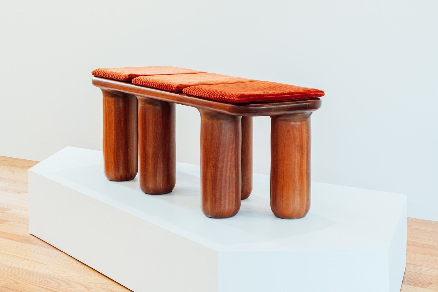 A piece of furniture designed by Michael Bennett, a wooden bench topped by velvet cushions and resting on a series of cylindrical wooden legs.