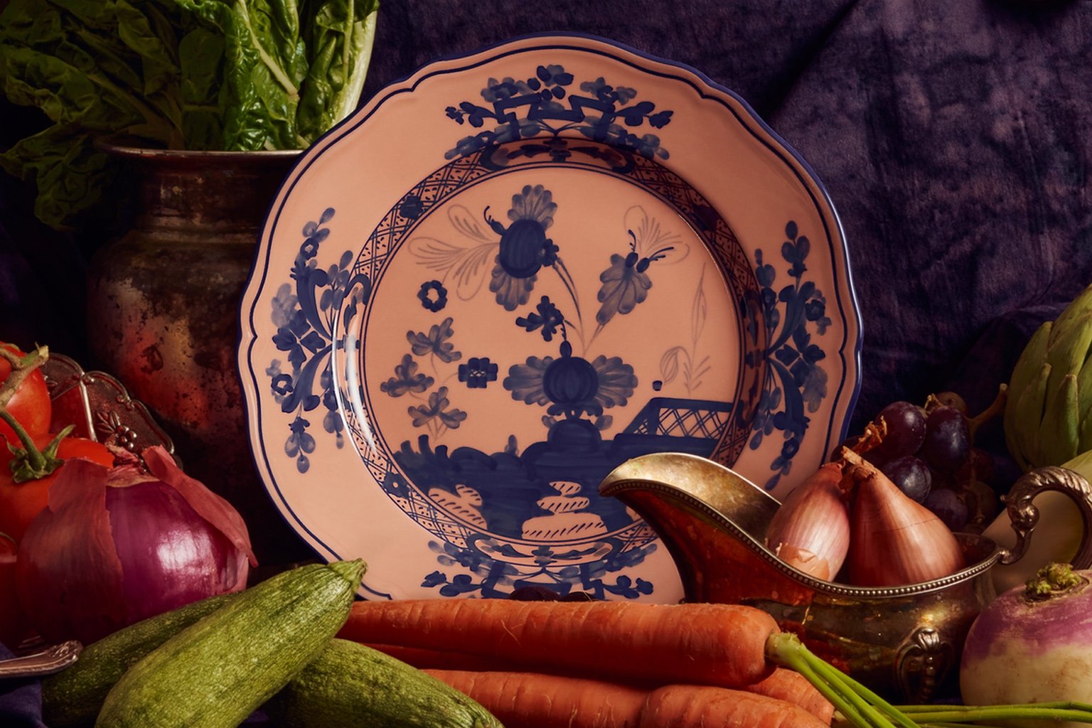 A cream-coloured dinner plate with an blue illustration of a garden. It's shown as part of a still life, surrounded by onions and carrots.