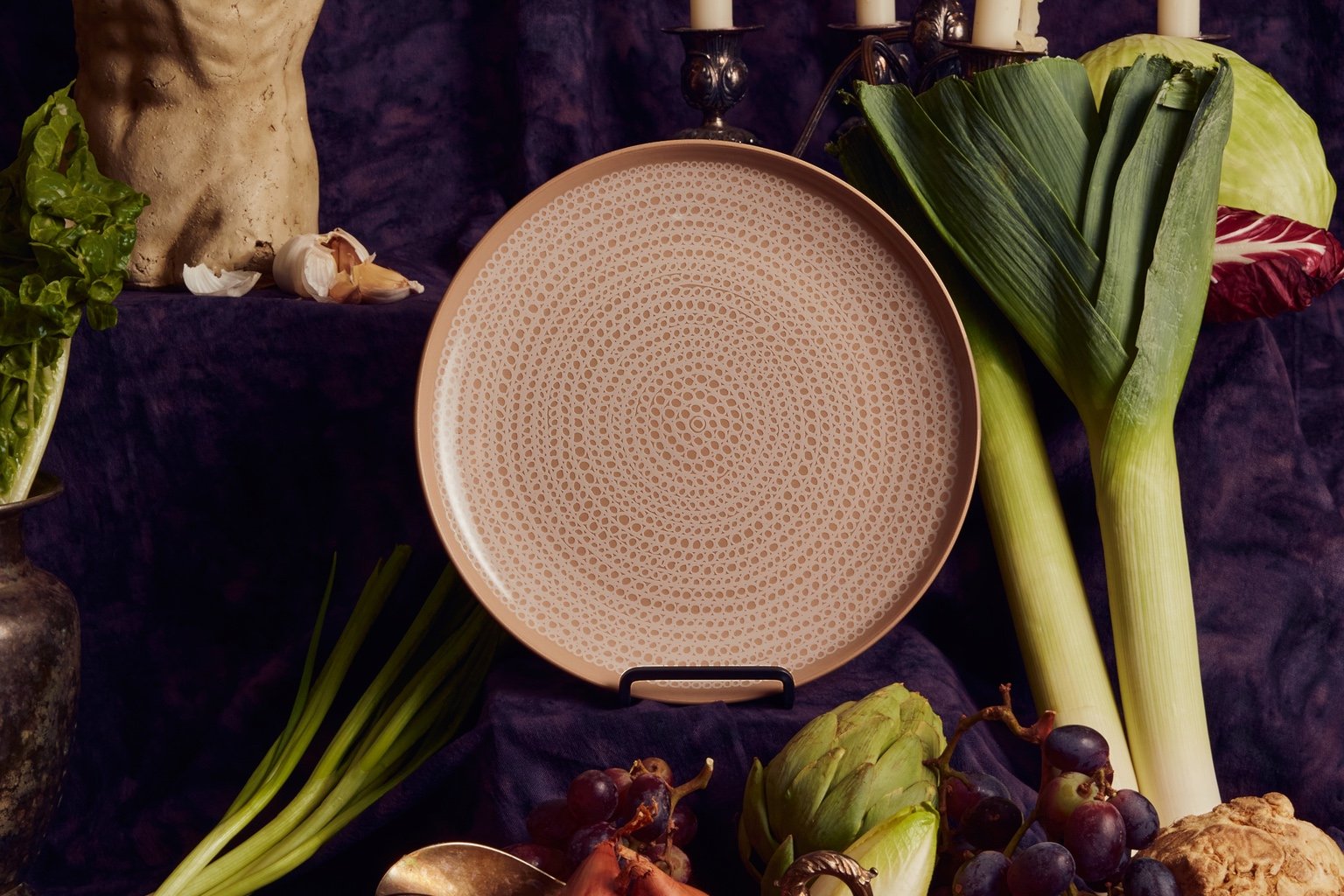 A beige dinner plate featuring a dotted geometric pattern. It's shown as part of a still life, with a small torso bust on one side and leeks on the other.