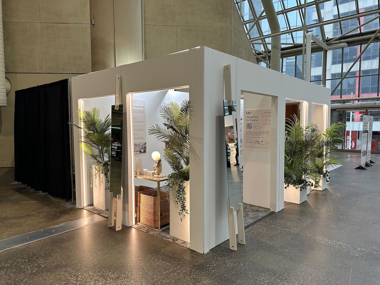 A white booth with large doorless openings, emerging ferns and standing mirrors balanced against outer walls for the LIV Student Design Challenge at IDS 2025.