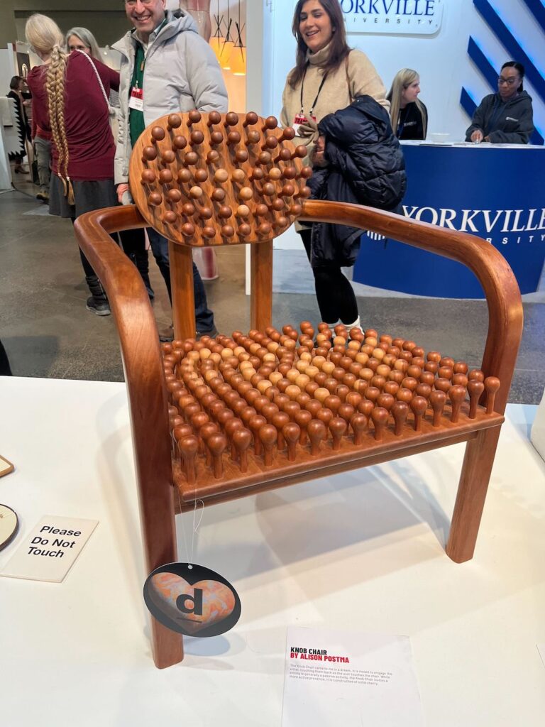 IDS 2025 display of Alison Potsma's Knob Chair, a cherry wood armchair covered in rounded knobs along its backrest and seat.