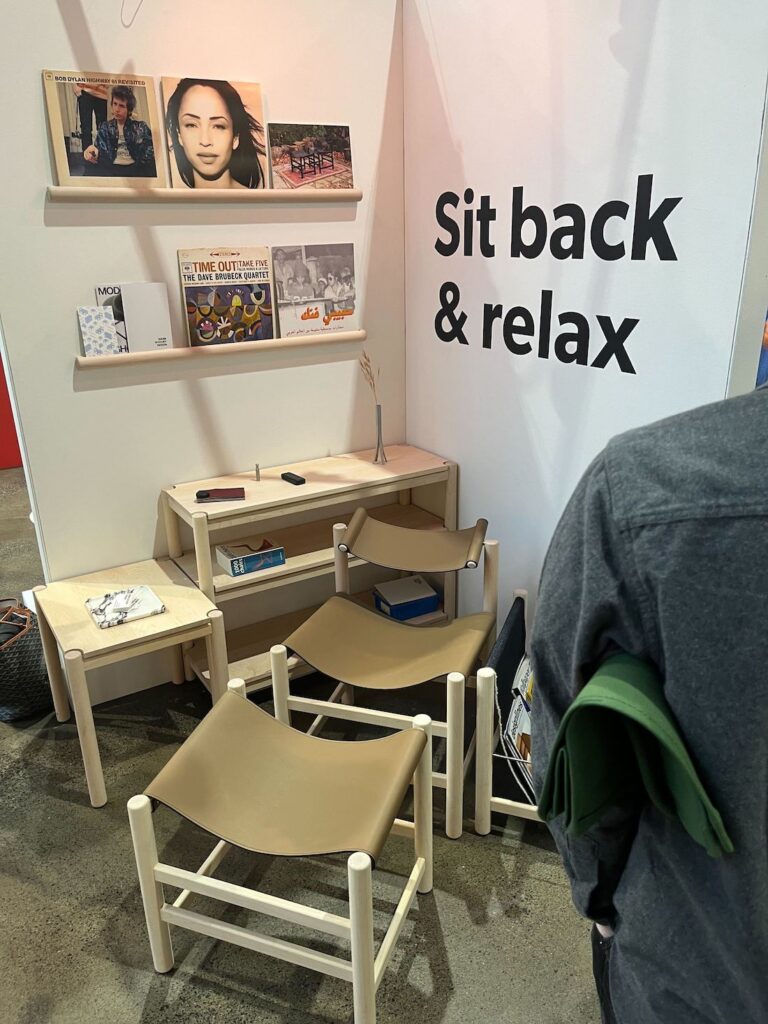 The Mark Khoury Design booth at IDS 2025, displaying three beige backless slung leather stools and a collection of wall-mounted shelves with records.