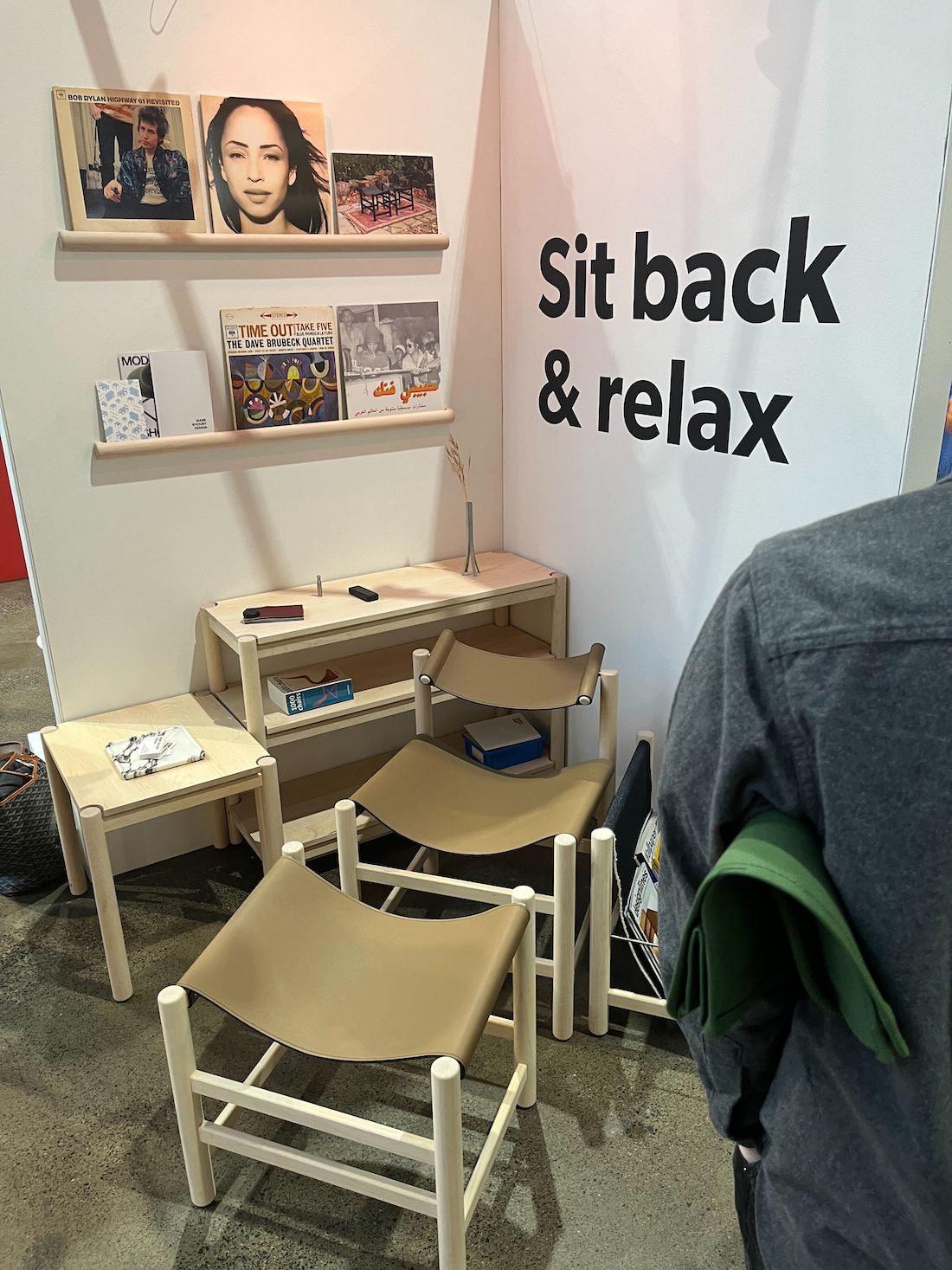 The Mark Khoury Design booth at IDS 2025, displaying three beige backless slung leather stools and a collection of wall-mounted shelves with records.