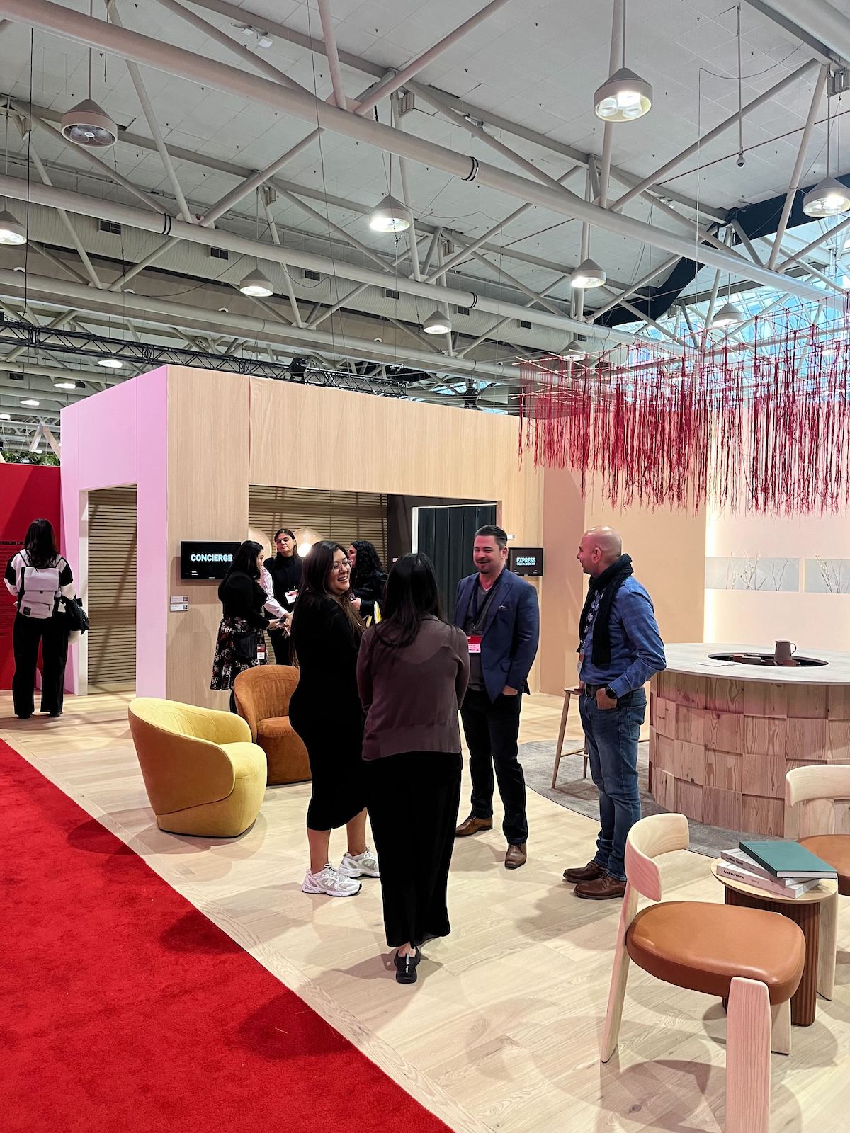 A reimagining of the hotel lobby space by SDI Design and Denizens of Design at IDS 2025, featuring a circular "hearth" and red ceiling installation.