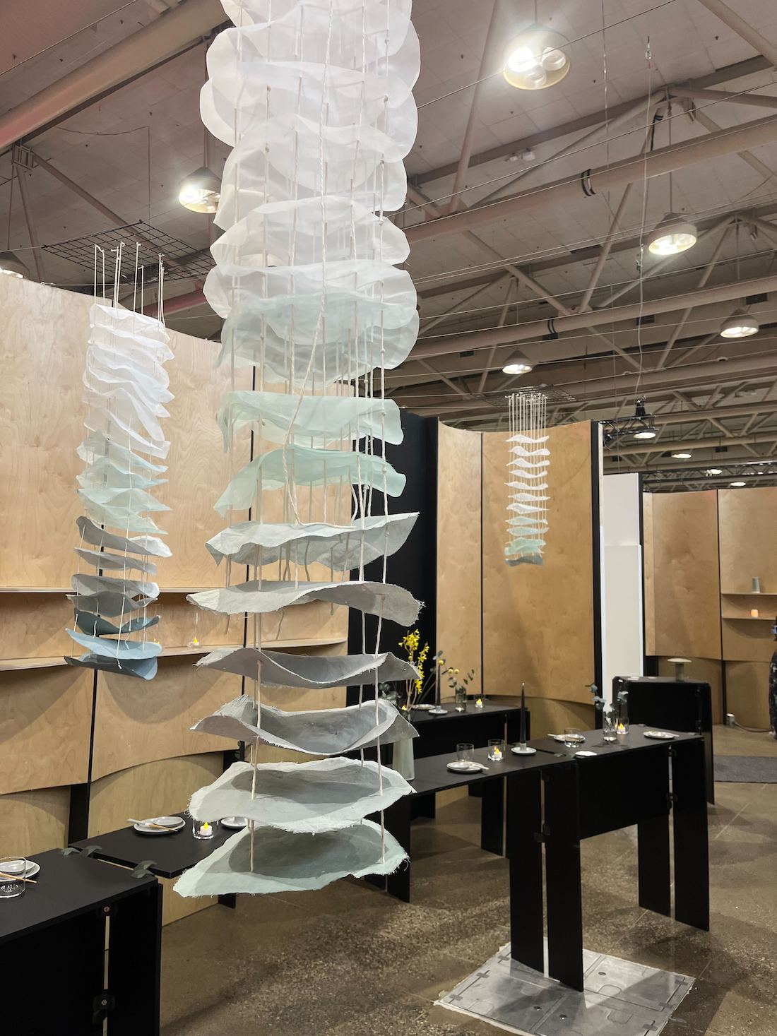 A redesigned hotel restaurant space by Waissbluth Architecture and AD Projects at IDS 2025, featuring long, turquoise and white hanging installations by Kristen Parry and black modular tables by PlayWood s.r.l..