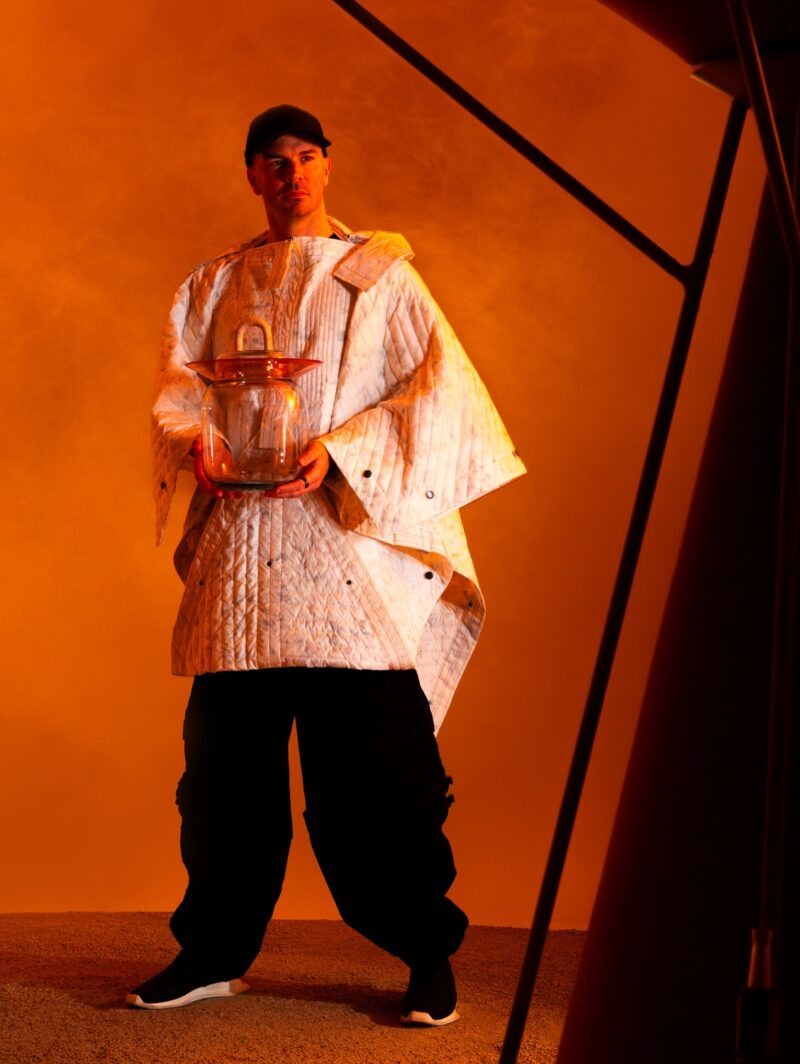Benjamin Hubert wears a white blanket-like jacket while holding a glass jar with a rounded handle.